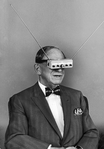 Erfinder Hugo Gernsback mit einer TV-Brille. Die „Brille“ ist ein Kasten mit Knöpfen und zwei absurd langen Antennen und ist mit einem Gurt vor die Augen gespannt, wie eine moderne VR-Brille.